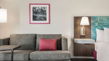 Une chambre d’hôtel moderne avec un canapé gris, un coussin rouge, une photographie encadrée accrochée au mur, une table de chevet, une lampe, un lavabo et un lit avec une literie blanche.