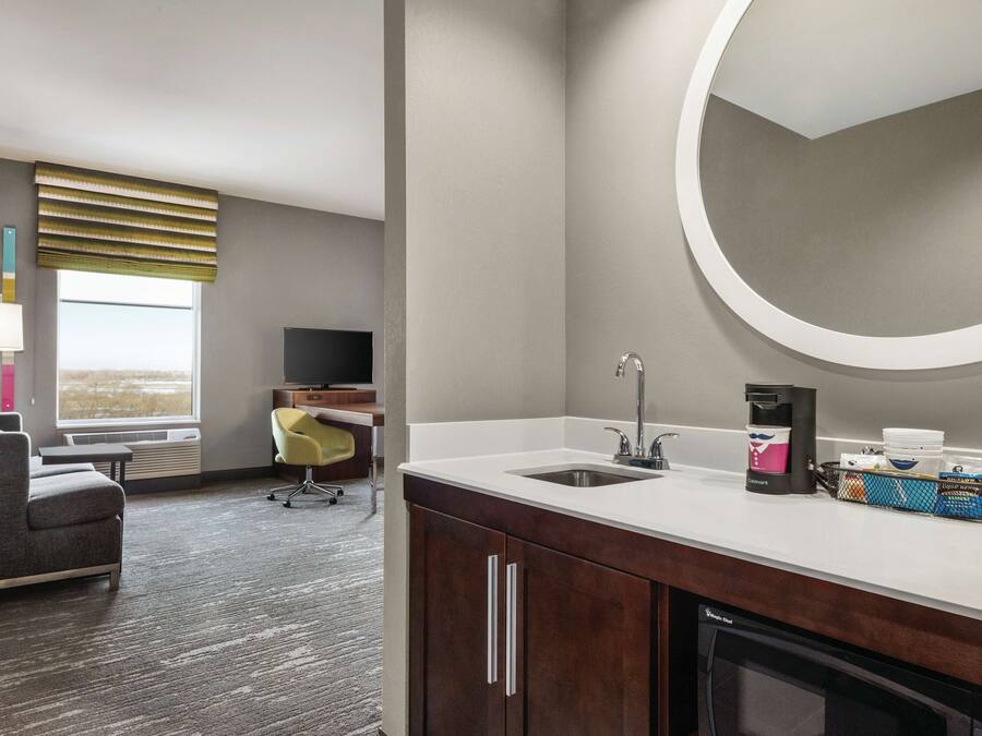 A modern hotel room with a kitchenette, a large window with blinds, a gray sofa, a small desk, and a flat-screen TV.