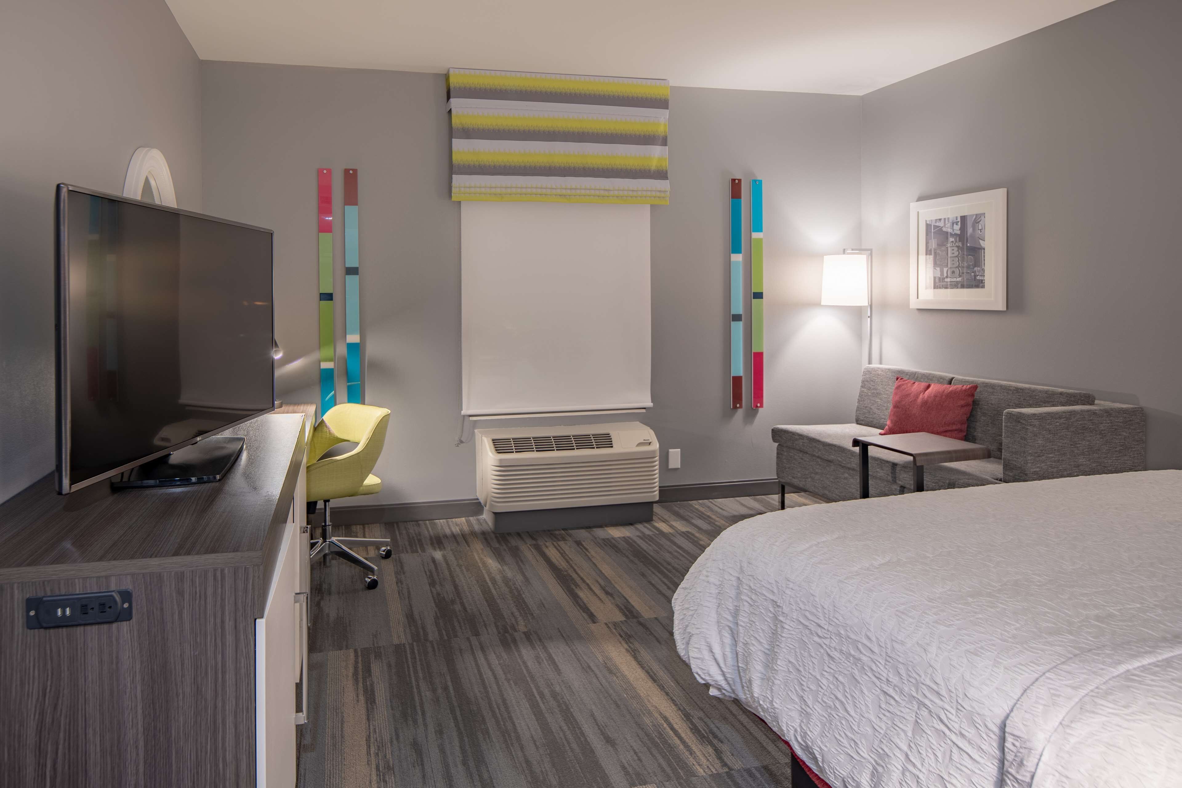 Une chambre d’hôtel moderne équipée d’un lit, d’un bureau avec une télévision, d’une chaise, d’un canapé et d’un climatiseur fixé au mur.