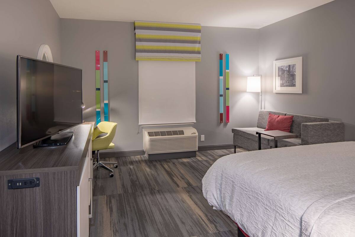 A modern hotel room with a bed, a desk with a TV, a chair, a sofa, and a wall-mounted air conditioner.