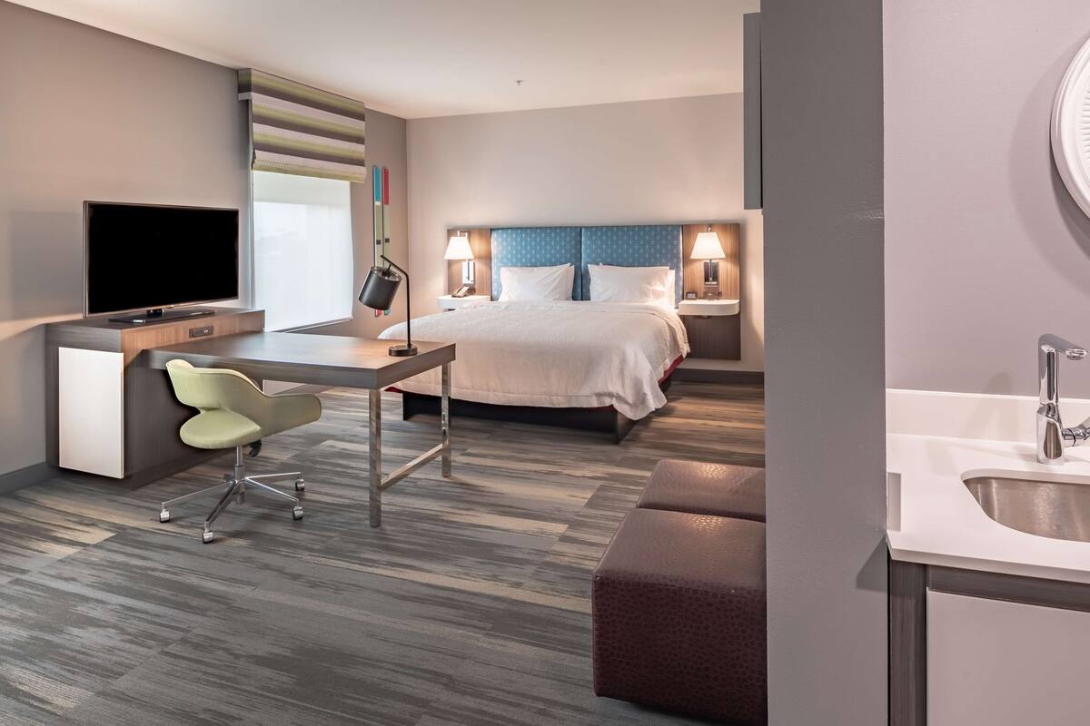 A modern hotel room with a bed, desk, chair, TV, and a bathroom with a sink and mirror.