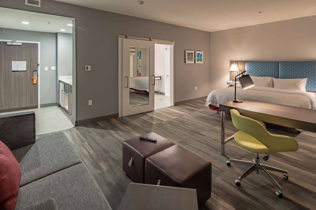 A hotel room with a bed, a desk, a chair, a small table, and a sofa. There is a door leading to a bathroom and a hallway.