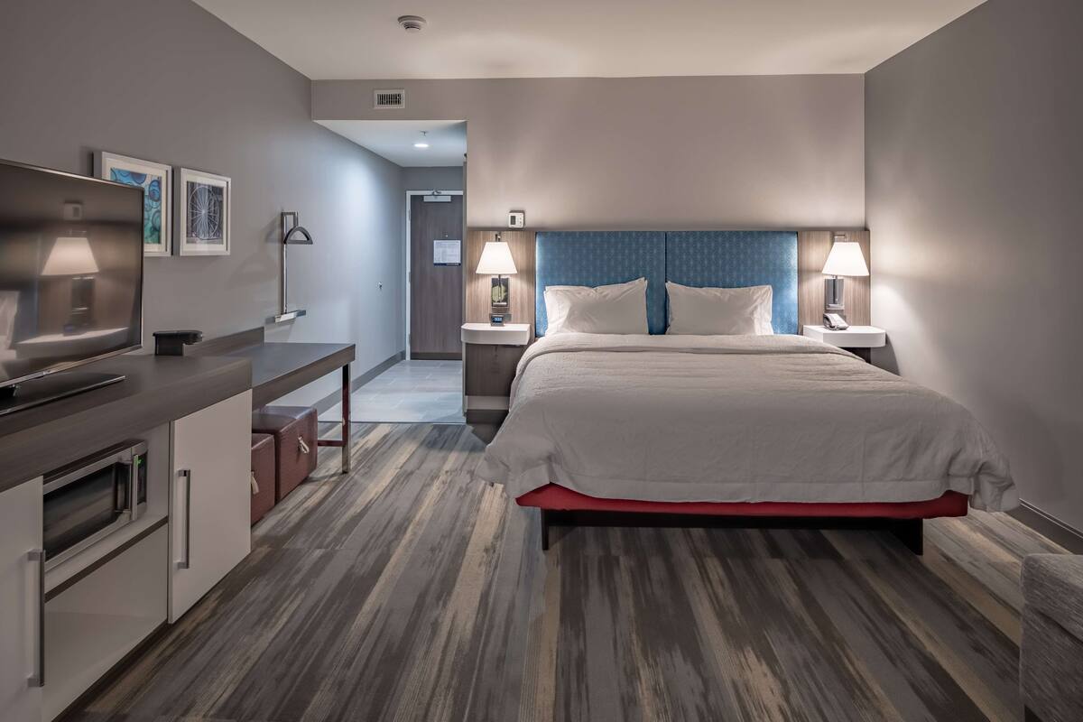 A modern hotel room with a large bed, bedside tables with lamps, a flat-screen TV, a kitchenette, and a view of the hallway.