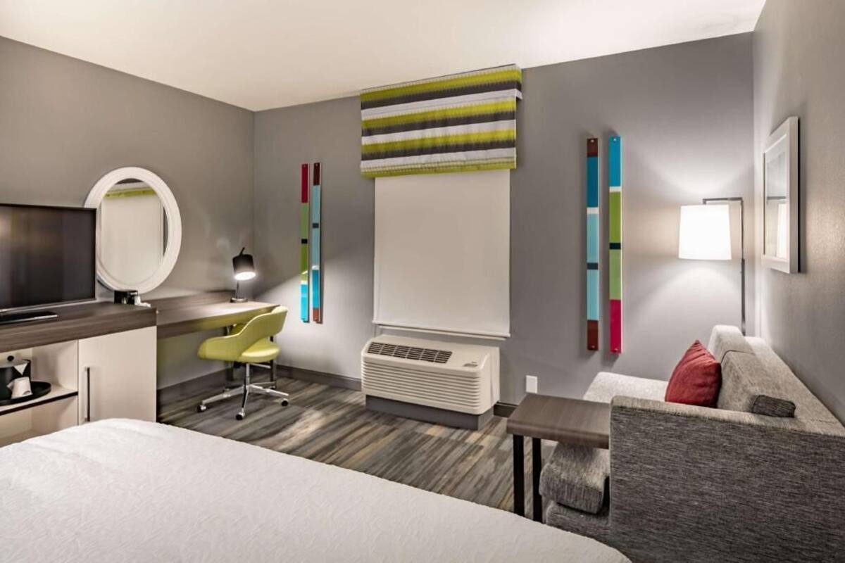 A hotel room with a bed, desk, chair, TV, air conditioning unit, and a sofa.