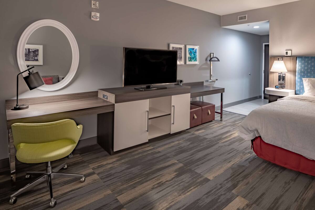 A hotel room with a bed, a desk with a flat-screen TV, a chair, a mirror, and a bedside table with a lamp.