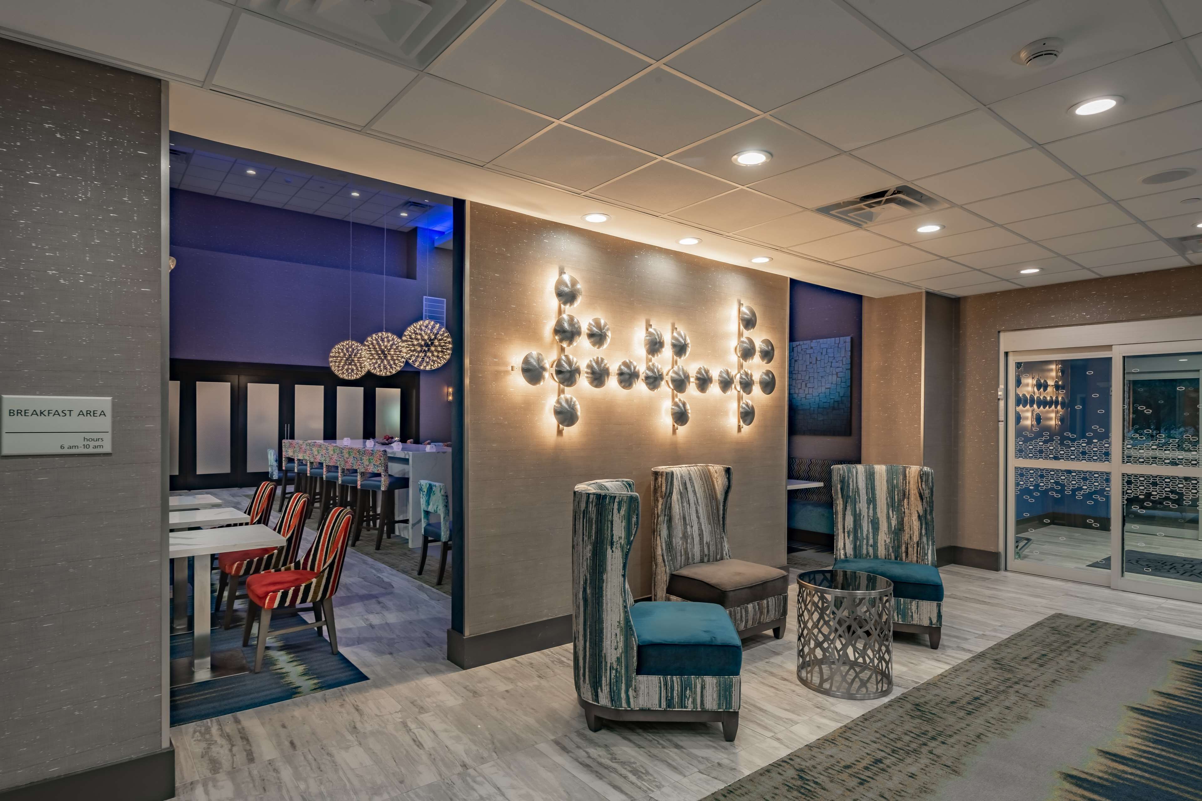 A modern hotel lobby with a breakfast area, seating with patterned upholstery, and a decorative wall with spherical elements.