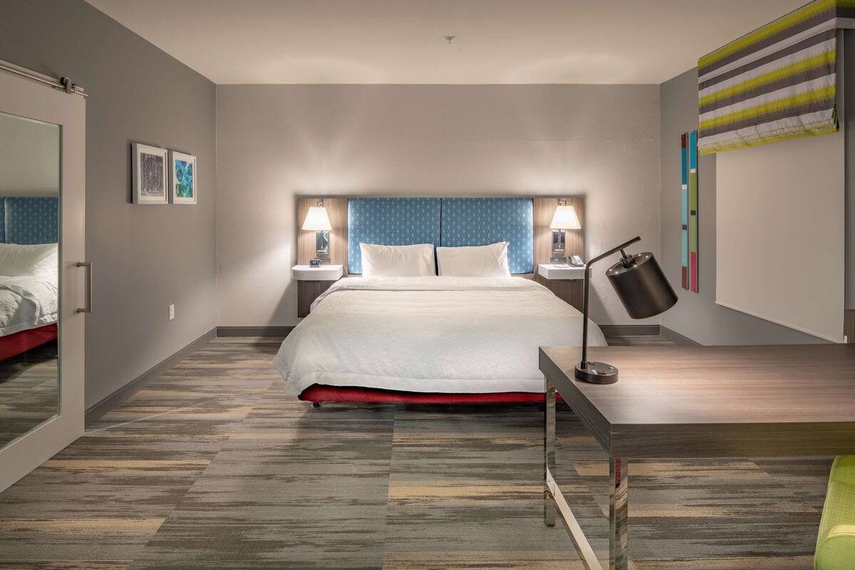 A modern hotel room with a large bed, two bedside tables with lamps, a desk with a lamp, and a mirror.