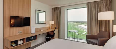 Habitación de hotel con una cama grande, un escritorio con televisor de pantalla plana, una mesita, una silla y vistas a un campo deportivo por la ventana.