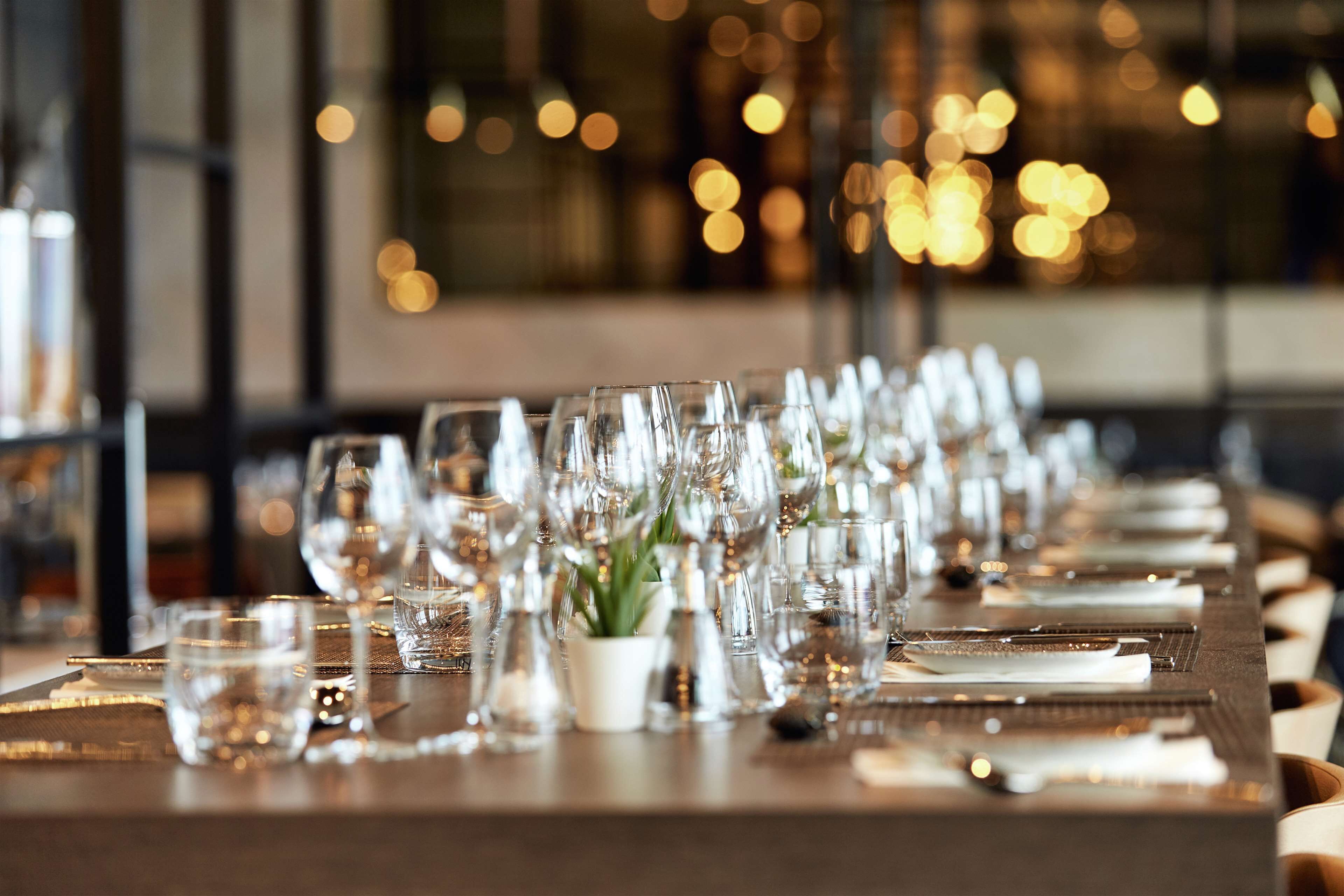 Una larga mesa de comedor dispuesta con múltiples copas de vino, platos y cubiertos, lo que sugiere un ambiente de cena formal.