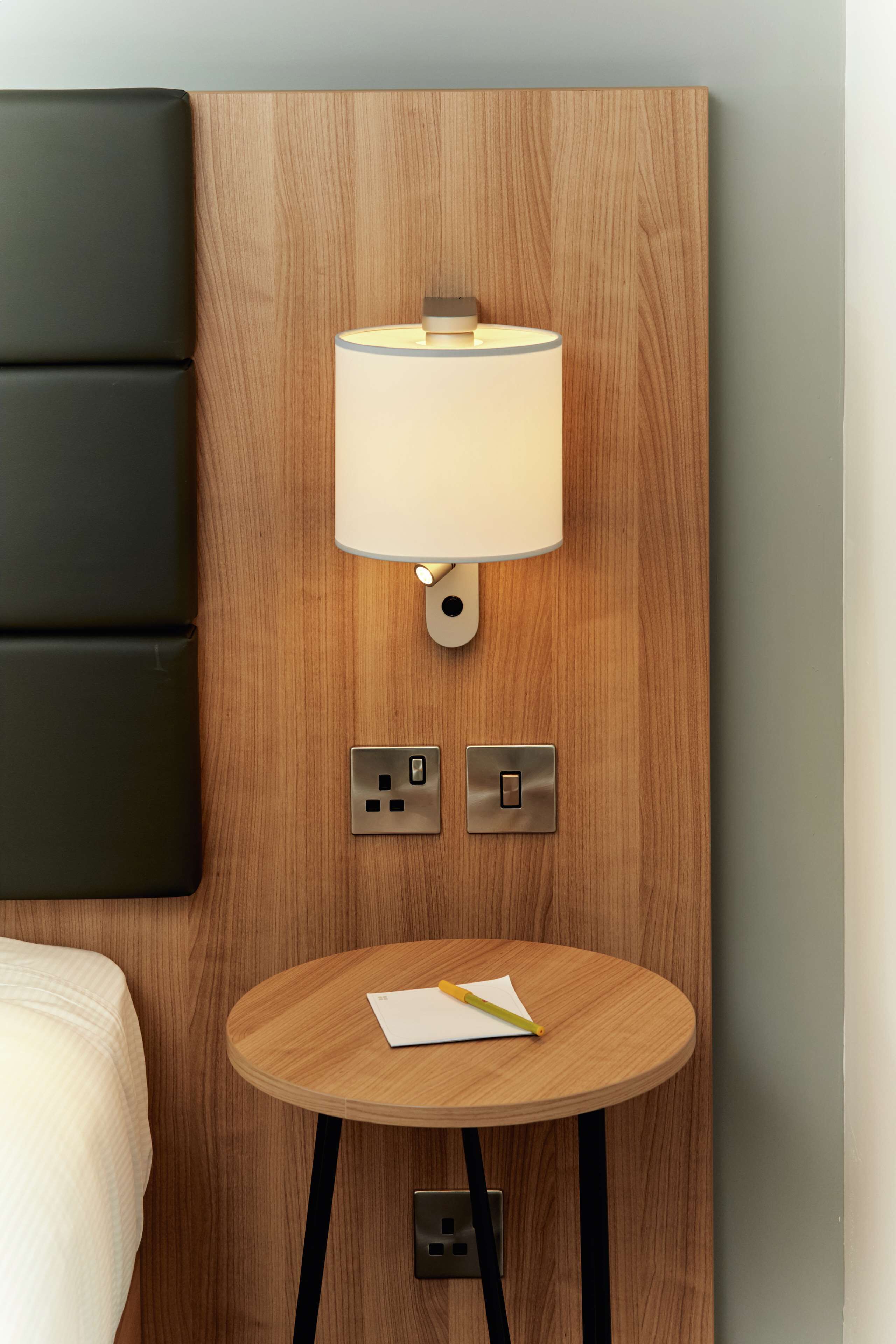 Een moderne kamer met een houten hoofdbord, een nachtkastje met een lamp, en een klein rond tafeltje met schrijfschrift en pen.