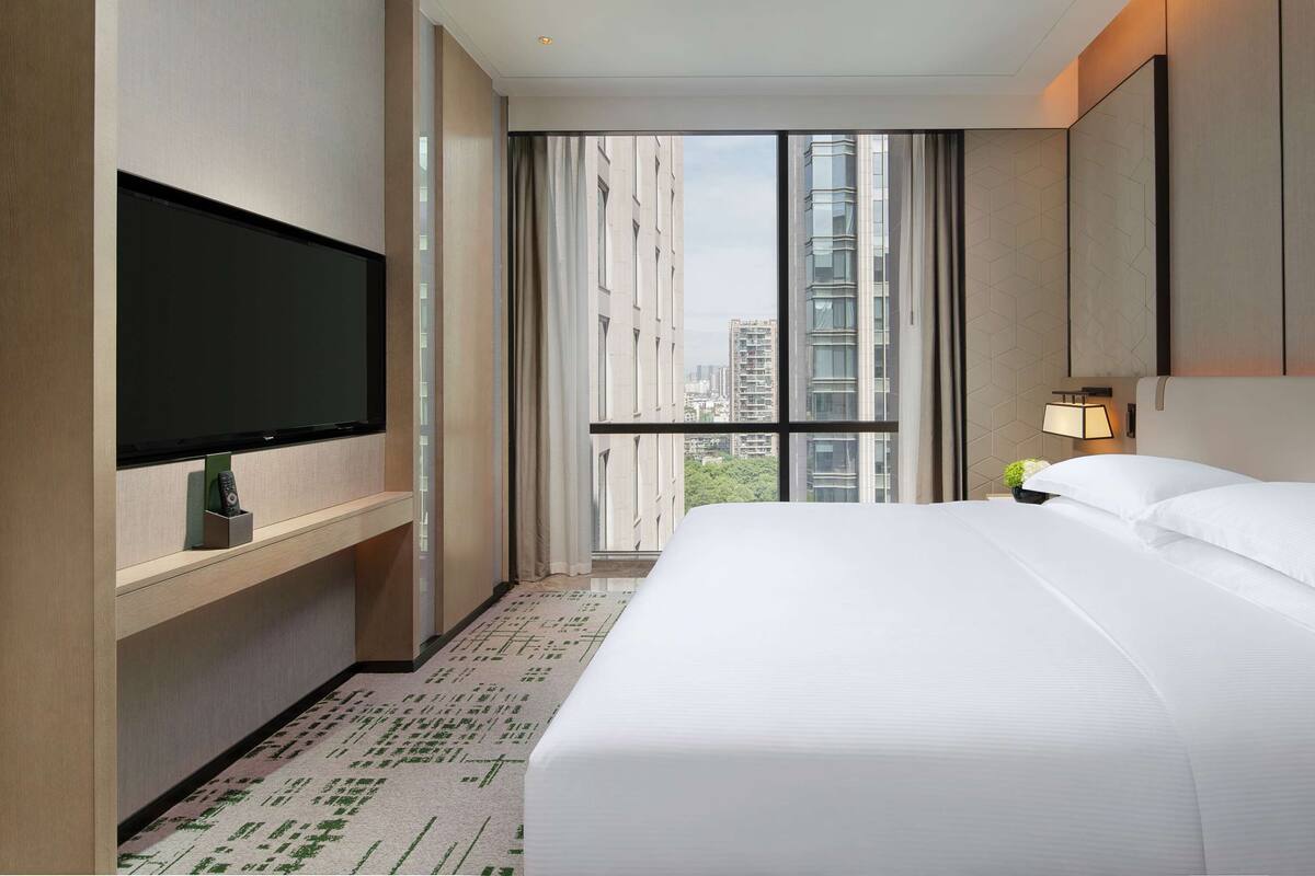 Habitación de hotel con una cama grande, televisión de pantalla plana, una mesita con lámpara y vista a la ciudad a través de un ventanal amplio.
