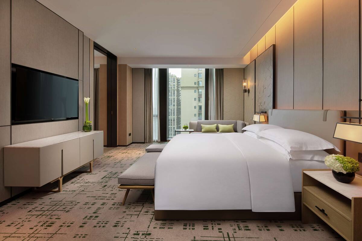 Una habitación de hotel moderna con una cama grande, un televisor de pantalla plana, una mesita de noche con lámpara y vistas a la ciudad a través de amplias ventanas.