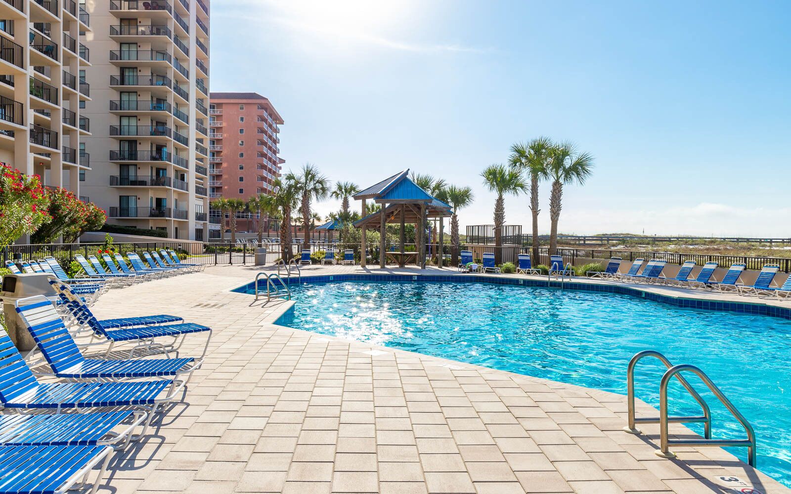 A swimming pool with lounge chairs and a gazebo, surrounded by palm trees and a high-rise building.