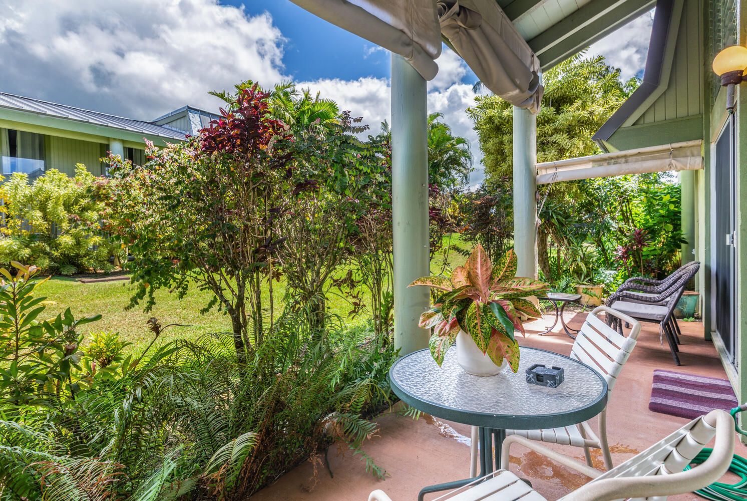 A covered outdoor seating area with a table and chairs, surrounded by lush greenery and a view of a building in the background.