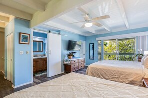 Une chambre avec deux lits, un ventilateur de plafond, une télévision, une salle de bain avec un lavabo et un miroir, et un balcon avec vue sur les arbres.
