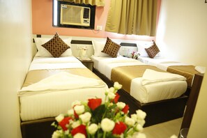 Habitación de hotel con tres camas, cada una con un diseño diferente de almohada. Se ve una ventana con cortinas, una unidad de aire acondicionado sobre la ventana y una mesita de noche con una lámpara y un florero.