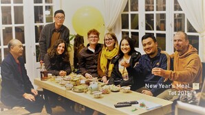 Een groep mensen zit rond een tafel met eten en drinken, in een ruimte met grote ramen en gordijnen.