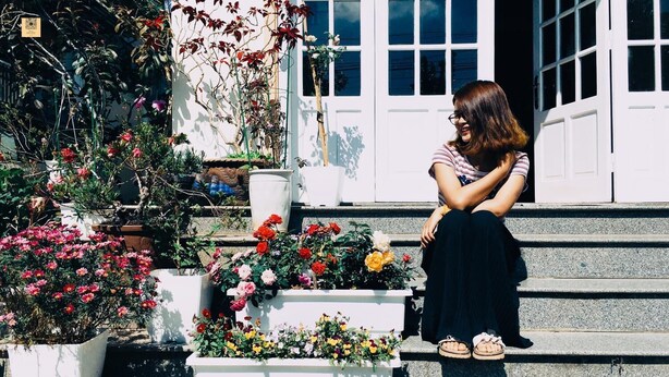 Een vrouw zittend op buitentrede, omringd door potplanten en bloemen.