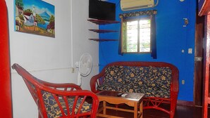 A room with a blue wall, a wooden sofa, a red chair, a small table, a TV mounted on the wall, and a window with a metal grill. - Rositas Hotel (San Juan del Sur)