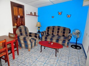 A living room with patterned sofas, a red coffee table, a wooden table, and a fan. - Rositas Hotel (San Juan del Sur)