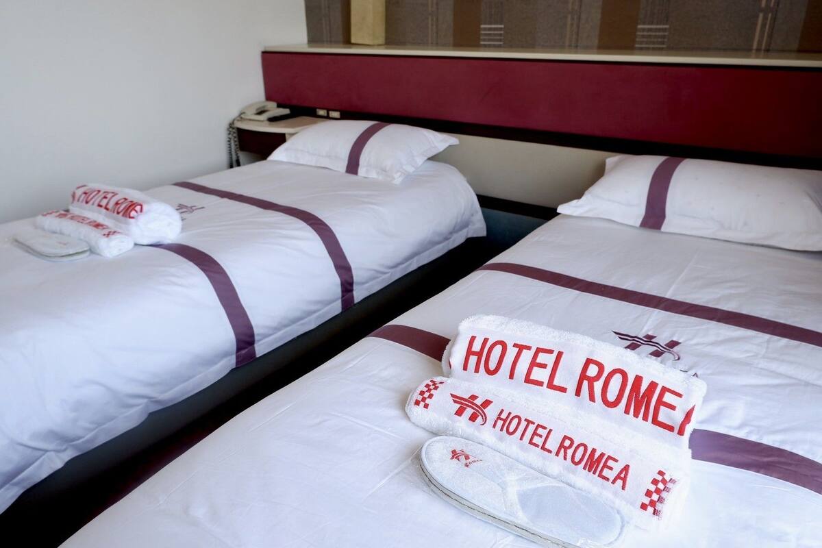 Ein Hotelzimmer mit zwei Betten, jedes mit einem Kopfkissen und einem Handtuch mit der Aufschrift 'HOTEL ROMA'. Auf dem Nachttisch steht ein Telefon.