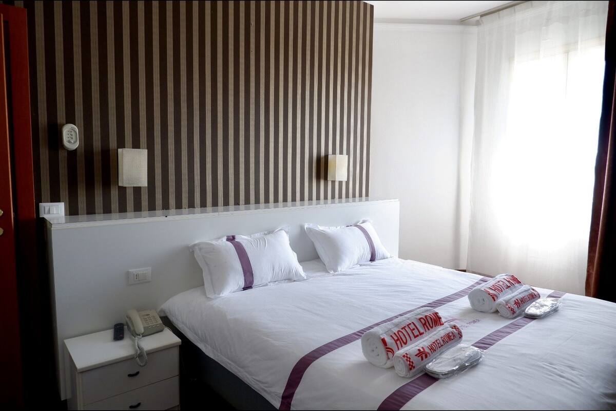 Ein Hotelzimmer mit einem großen Bett, weißer Bettwäsche und einem gestreiften Kopfteil. Ein Nachttisch mit Telefon und Lampe ist vorhanden.