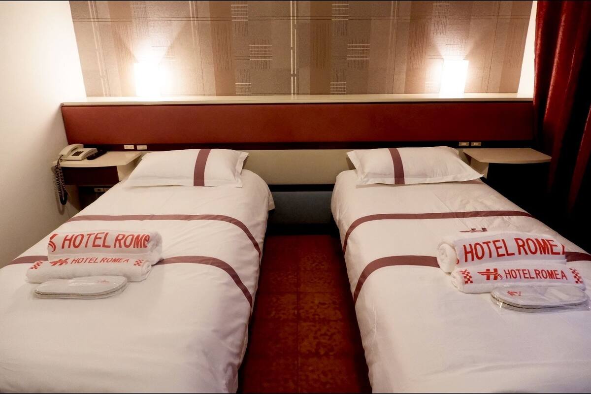 Ein Hotelzimmer mit zwei Betten, jedes mit einem Kopfkissen und einem Handtuch, versehen mit dem Schriftzug 'HOTEL ROMA'.