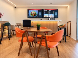 A modern dining area with a wooden table and orange chairs, a laptop on a desk, and a menu display above the counter. - K2 Guest House (Singapore)