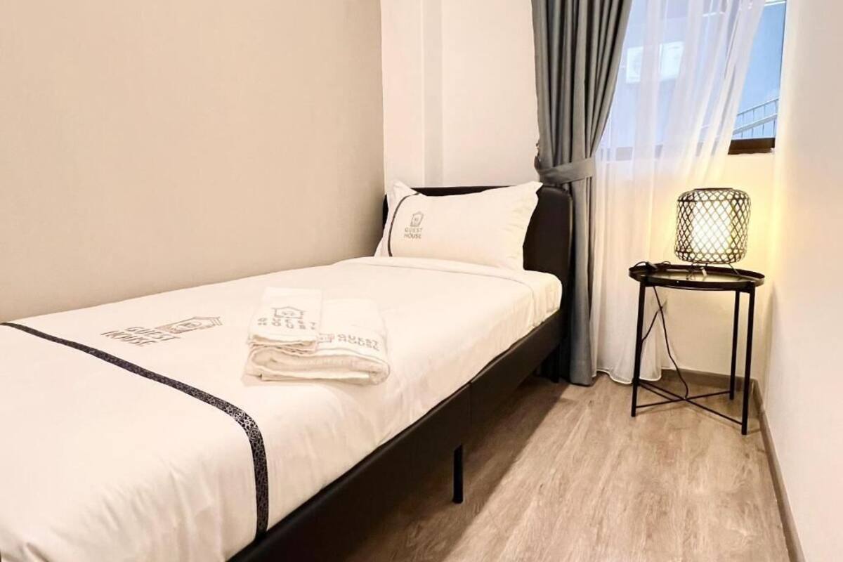 A small, neatly made bed with a white sheet and a pillow, a small table with a lamp, and a window with curtains.