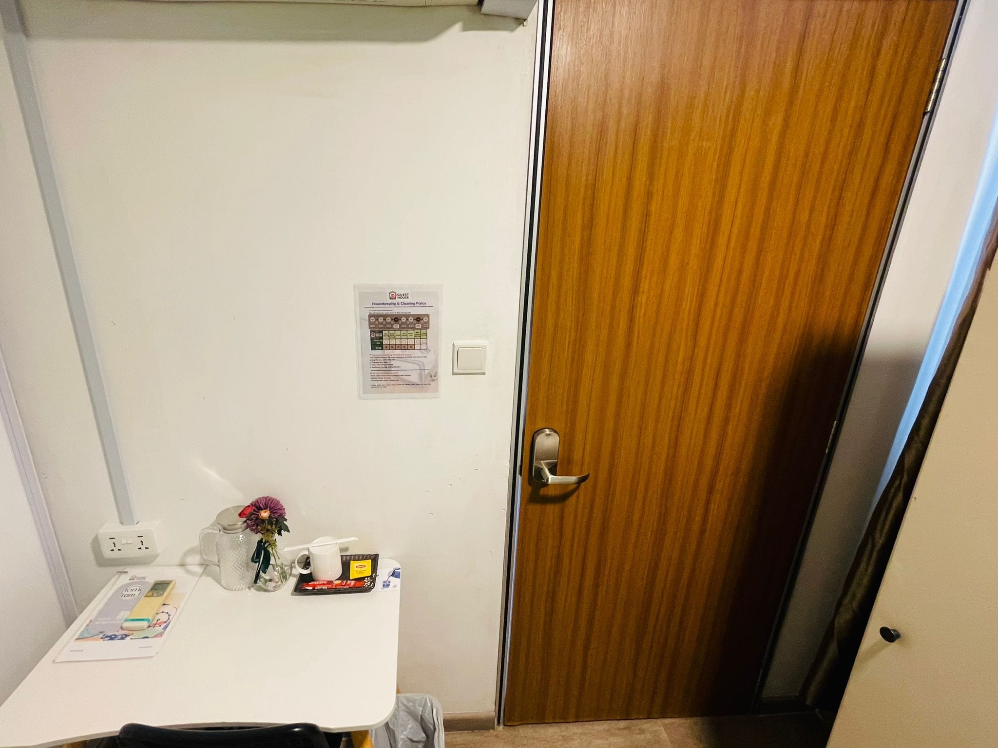 Una habitación con una mesa blanca, una puerta de madera, un interruptor de luz y un aviso en la pared.