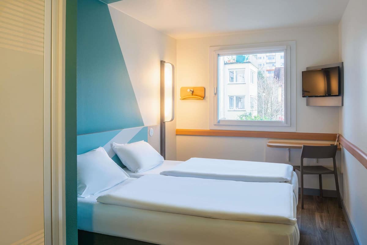 A hotel room with two beds, a desk, a chair, a TV, and a window with a view of buildings.