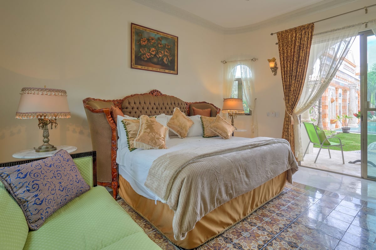 Un dormitorio con una cama grande, una mesita de noche, una lámpara, un sillón verde y vistas al exterior.