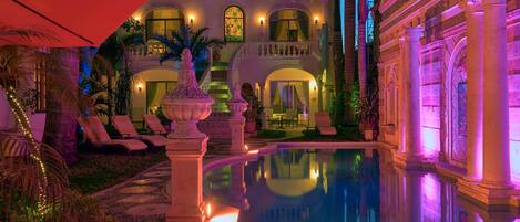 Un área de piscina lujosa con una fuente, sillas de descanso y un edificio imponente con ventanas de arco y columnas.
