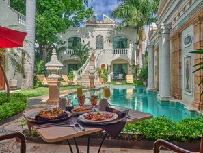 Un comedor exterior de lujo con piscina, rodeado de arquitectura ornamentada y plantas tropicales.