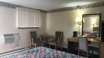 A hotel room with a bed, two chairs, a small table, a desk, a mirror, and an air conditioning unit.