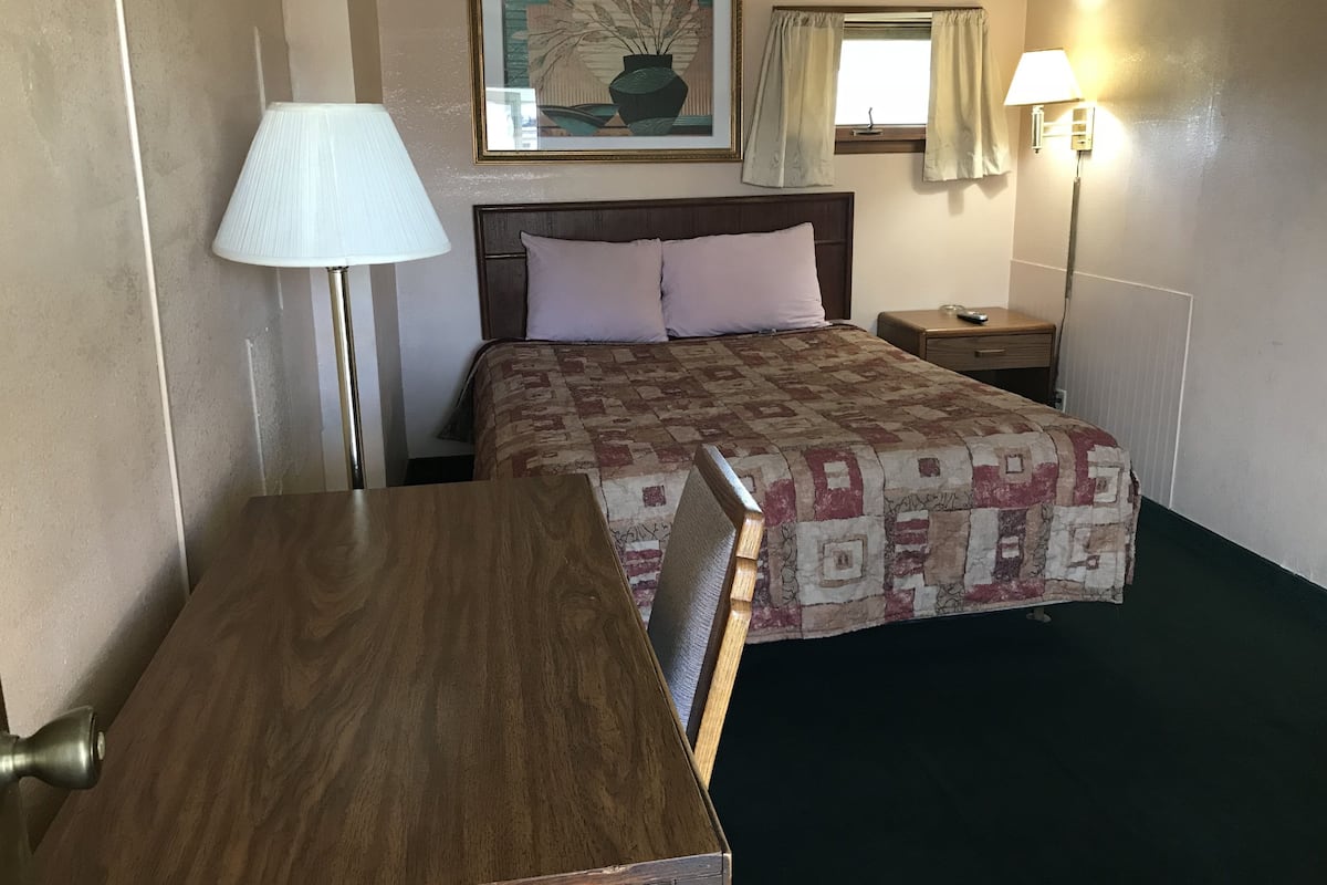 Hotellihuone, jossa on puinen sänky, yöpöytä, lamppu, seinällä oleva kehystetty kuva ja ikkuna, jossa on verhot.