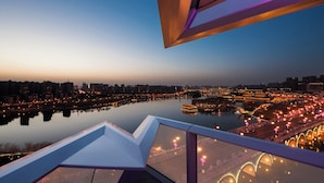 A panoramic view of a cityscape at dusk with a river in the foreground, illuminated buildings, and a modern bridge.