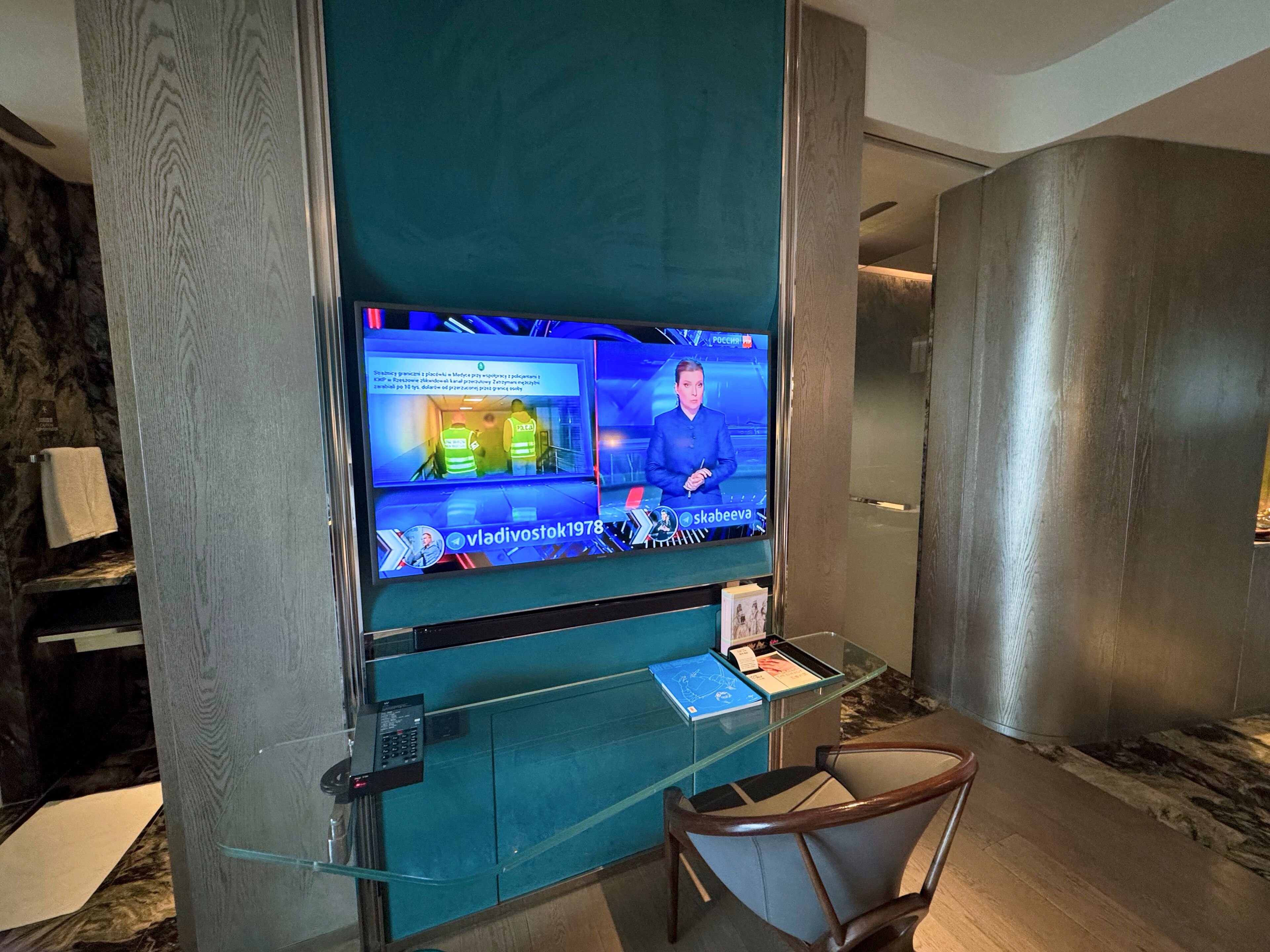 A modern hotel lobby with a large flat-screen TV displaying a news channel, a glass desk with a chair, and a marble floor.