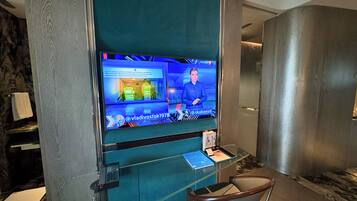 A modern hotel lobby with a large flat-screen TV displaying a news channel, a glass desk with a chair, and a marble floor.