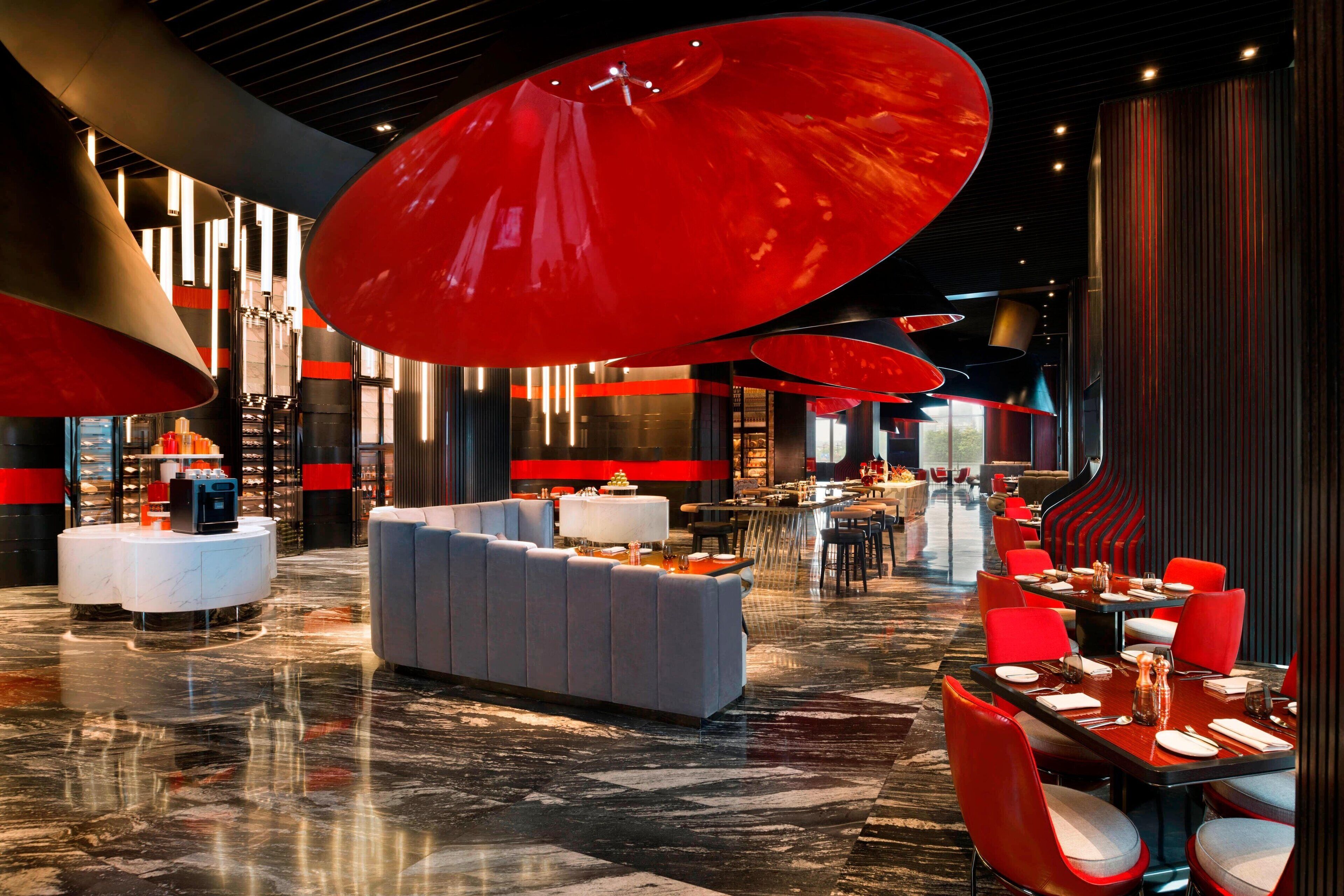 A modern hotel lobby with a marble floor, red and grey seating areas, and large red pendant lights.