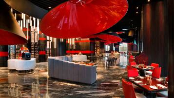 Un moderno vestíbulo de hotel con suelo de mármol, zonas de estar en rojo y gris, y grandes lámparas colgantes rojas.
