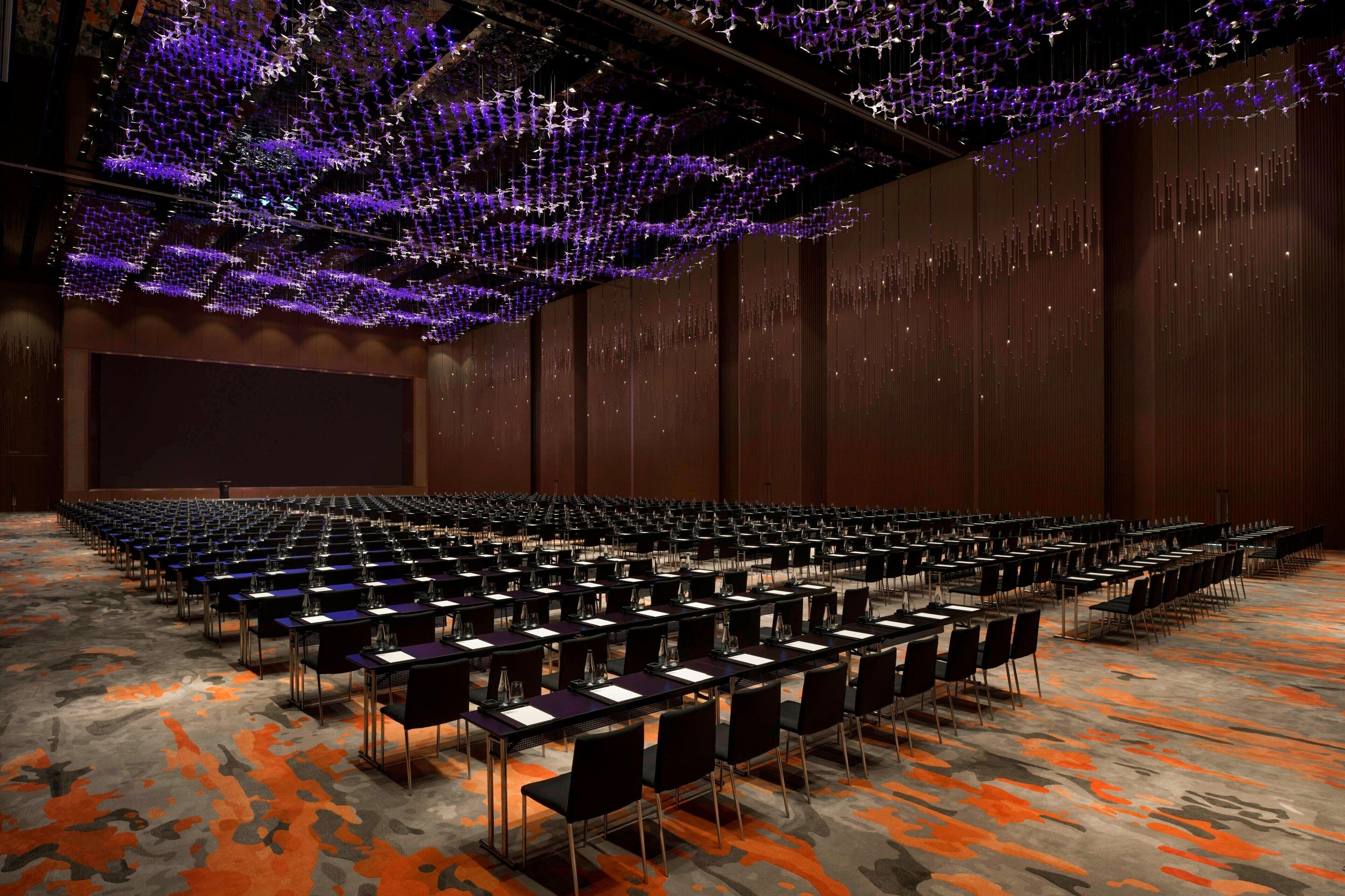 A large conference room with rows of tables and chairs, a large screen, and a ceiling with hanging lights.
