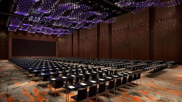A large conference room with rows of tables and chairs, a large screen, and a ceiling with hanging lights.