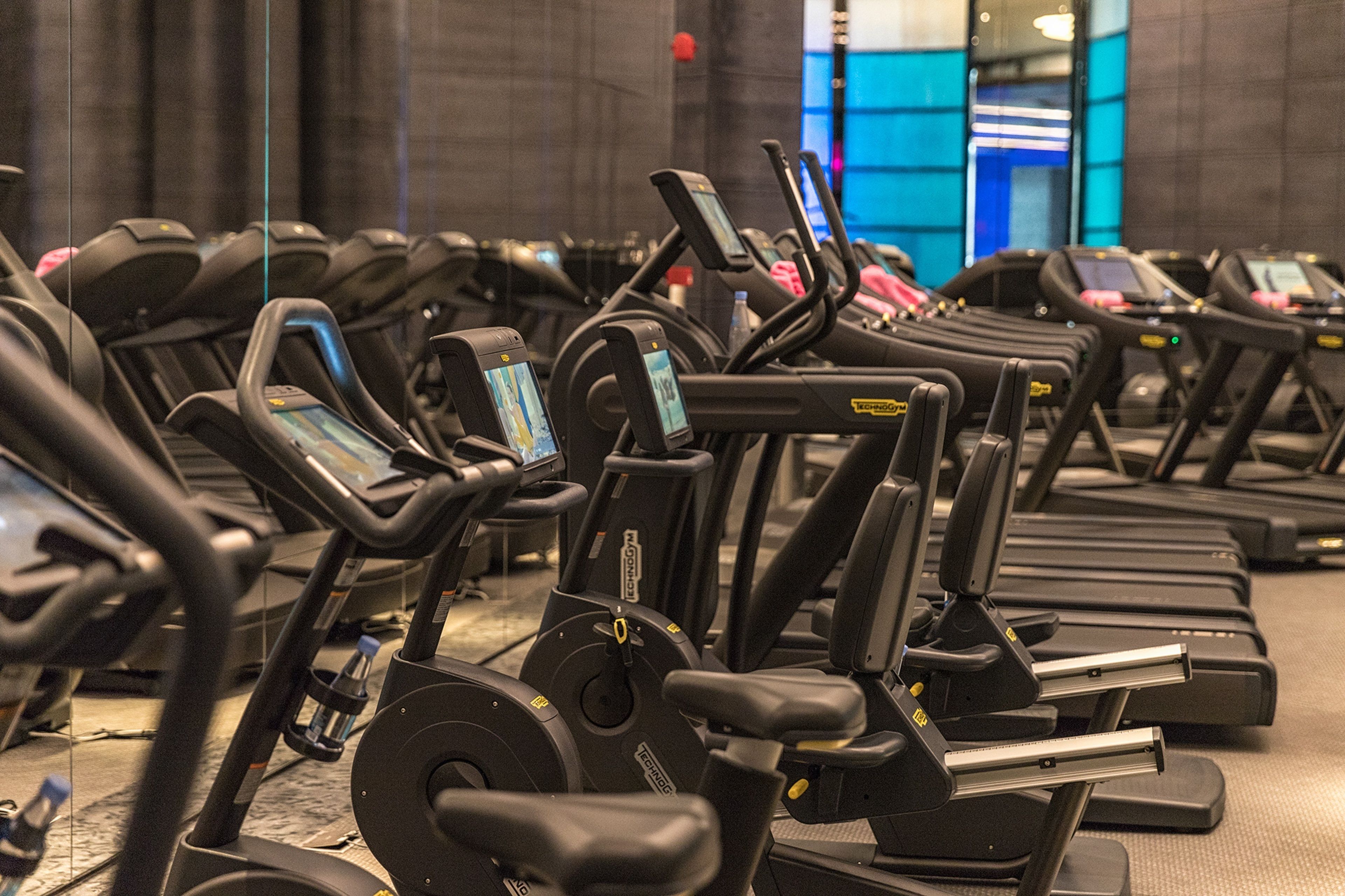 Varias bicicletas estáticas en un gimnasio con un espejo grande y un fondo iluminado en azul.
