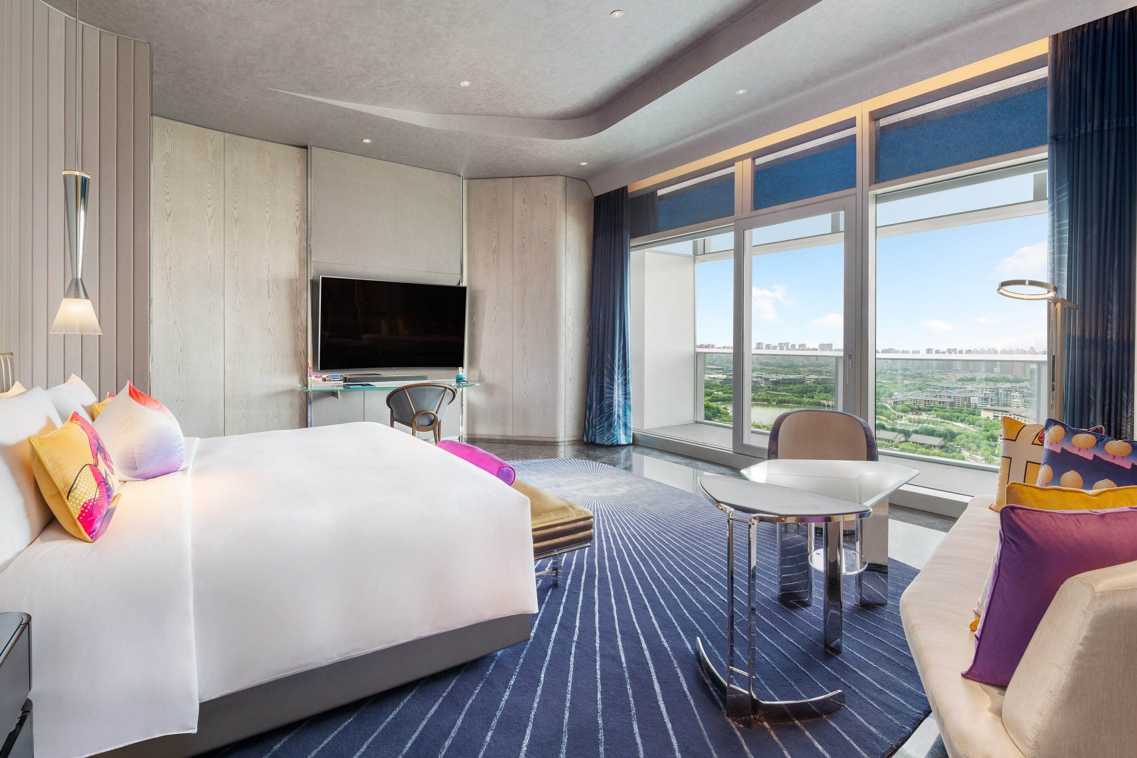 A modern hotel room with a large bed, a TV, a dining area, and a view of the city.