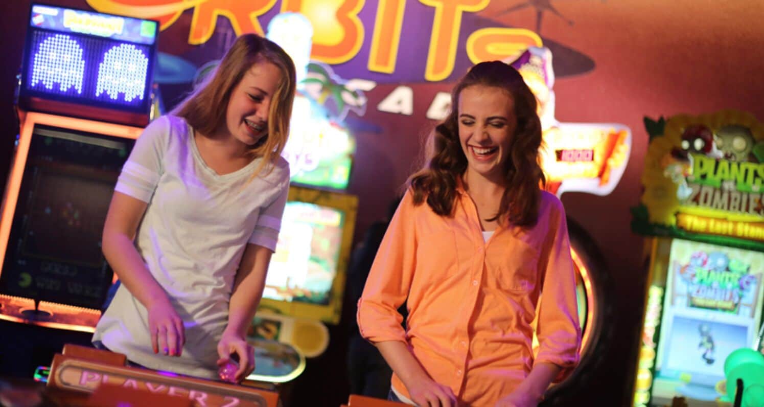 Twee personen spelen een spel in een fel verlichte speelhal met neonreclames en diverse spelautomaten.