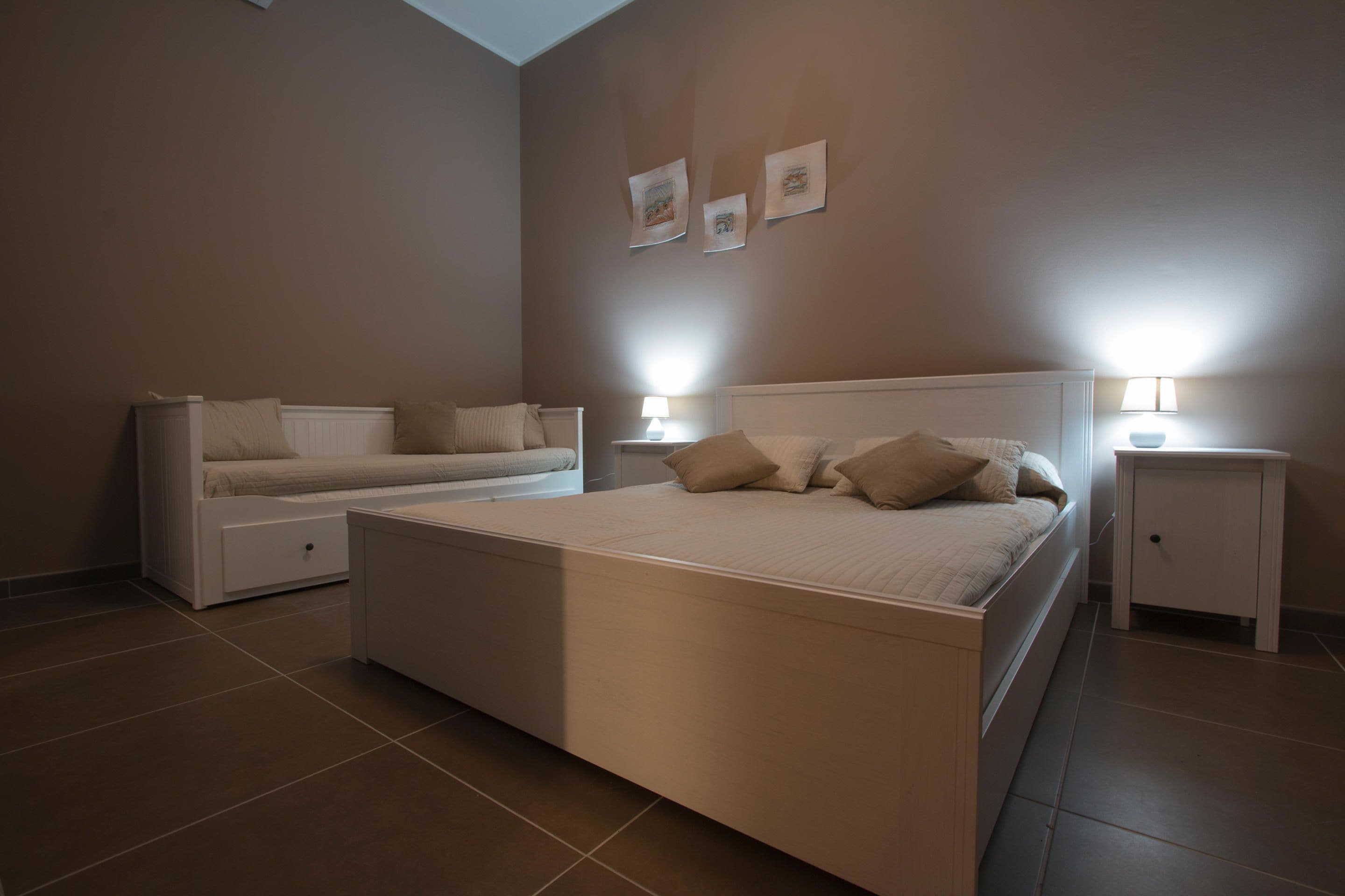 Un dormitorio con una cama grande, un sofá cama más pequeño, dos mesitas de noche con lámparas y tres cuadros en la pared.