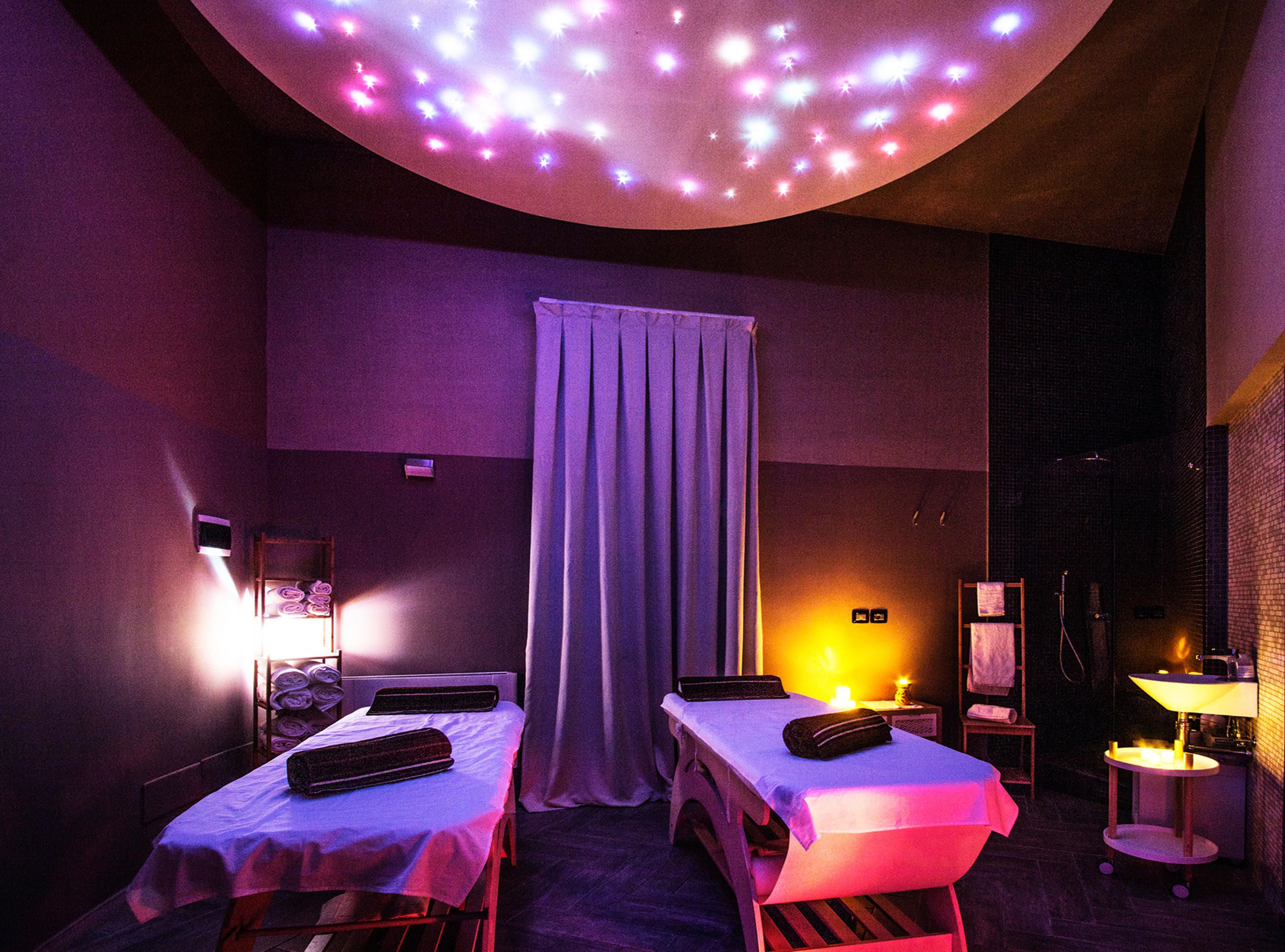 Une pièce avec deux lits, une table de massage, une petite table avec une lampe et un luminaire au plafond.
