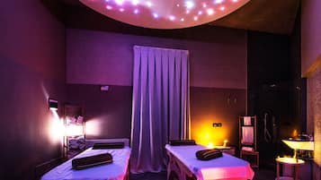 Une pièce avec deux lits, une table de massage, une petite table avec une lampe et un luminaire au plafond.