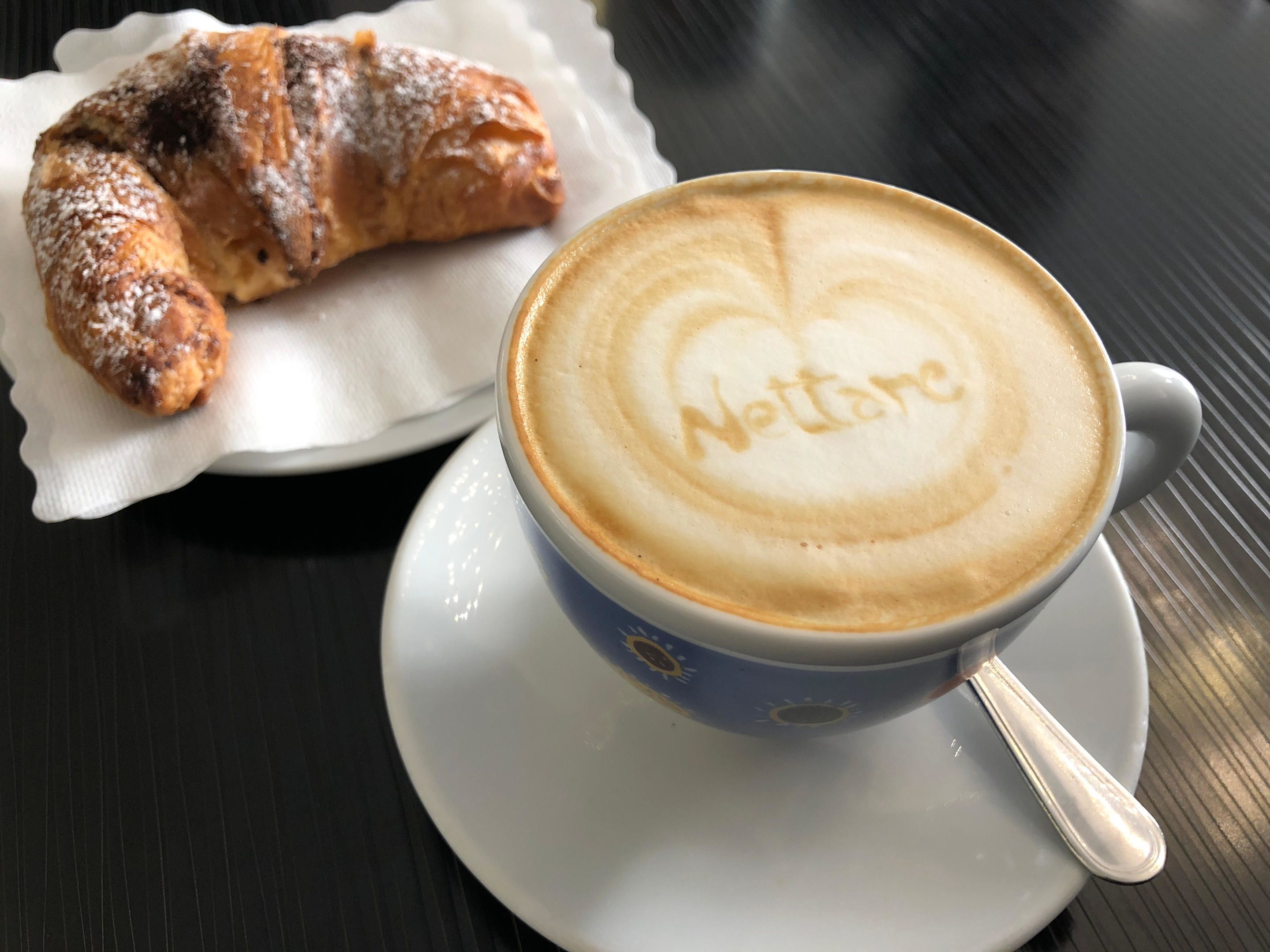 A cup of coffee with a heart-shaped latte art and a croissant on a white plate.